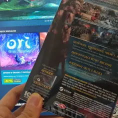 Dłoń trzyma pudełko gry z widokiem na ekran komputera ze sklepem Steam.