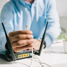 Osoba w niebieskiej koszuli trzyma wtyczkę kabla sieciowego przy routerze Wi-Fi.