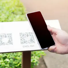 Dłoń trzyma smartfon nad tabliczką z kodami QR i napisem "ENGLISH".