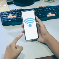 Dłoń trzyma telefon z ikoną Wi-Fi na ekranie, na biurku z klawiaturą.